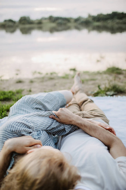 Young man hugs of a beautiful woman. Close-up hands together. Lovers lie on a blanket near the river - Фото, изображение