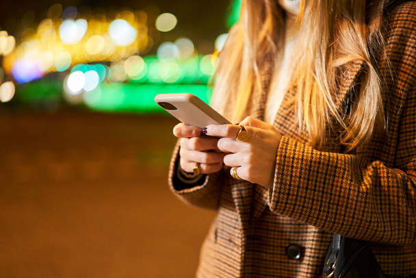 Primer plano de una joven rubia usando un teléfono inteligente, escribiendo un mensaje, en una ciudad por la noche, con retroiluminación y tráfico. - Foto, Imagen