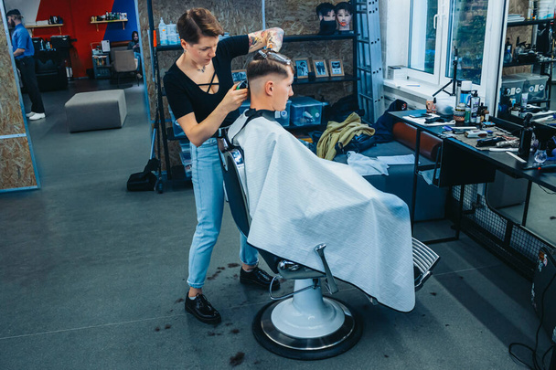 MINSK, BELARUS - OCTOBER 14, 2020: A tattooed woman hairdresser at a barbershop cuts a young guy's hair for a short haircut. - Foto, afbeelding