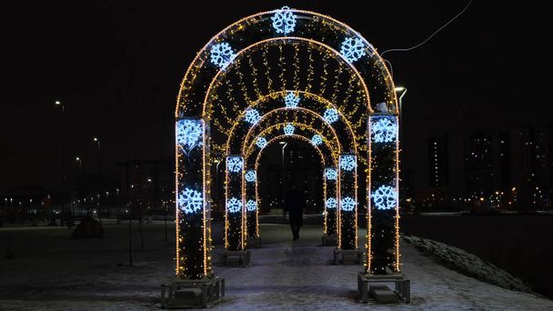 Ano Novo Decoração luminosa de Natal no parque da cidade no fundo do céu preto. Conceito. Um homem que passa pelo túnel luminoso com guirlandas douradas e flocos de neve azuis.  - Foto, Imagem