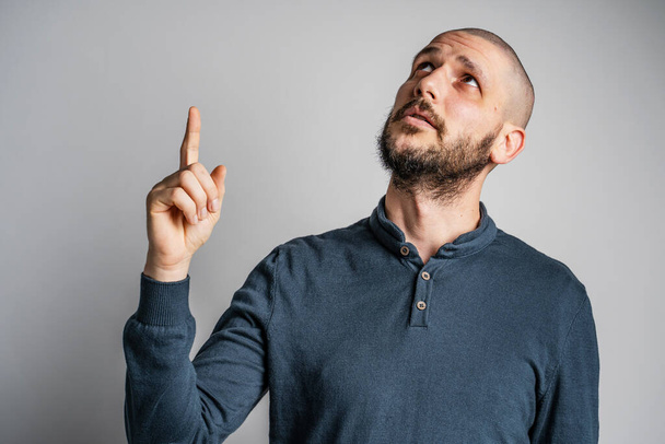 Front view portrait of adult caucasian man with beard and short hair pointing up while standing in front of white wall thinking with copy space studio shot - Photo, image