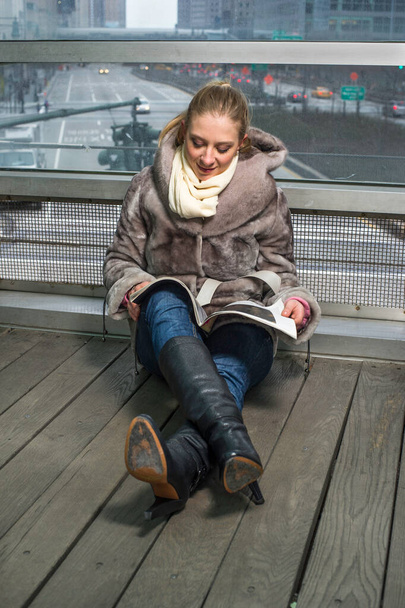 A pretty young woman is sitting on the floor by a big glass wall, reading and smiling. The background is a city street scene - Foto, imagen