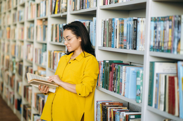 Stylish attractive female student in the library, standing near bookshelf. Beautiful caucasian girl in eyeglasses reading a book, learning some information for exams, education concept - Photo, image