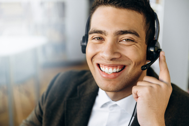 Portrait of hispanic operator of call center. Satisfied male consultant sitting at the desk, wearing headset looks directly at the camera with friendly smile - 写真・画像