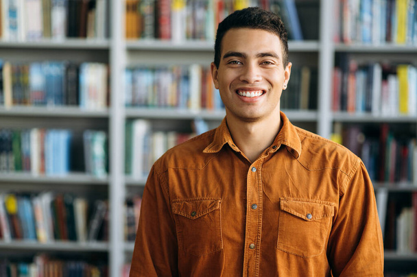 Portrait of successful hispanic business man looks directly at the camera, smile. Happy male freelancer or student standing in modern office or university library - Photo, Image