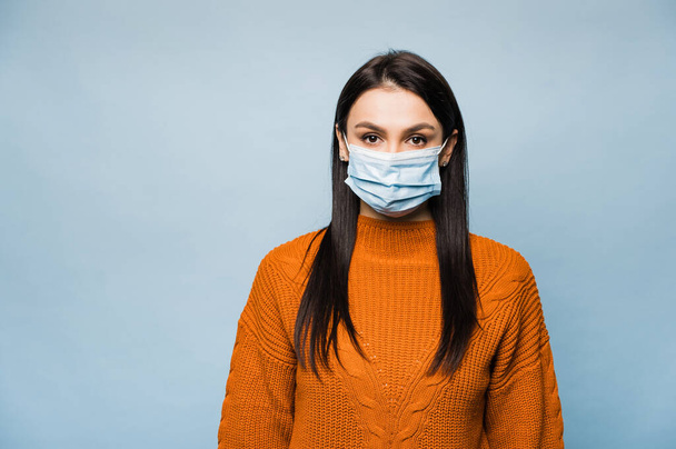 Portrét bělošské brunetky v oranžovém svetru, se sterilní obličejovou maskou z koronaviru covid-19 během pandemické karantény, se dívá přímo do kamery, stojí na izolované modré - Fotografie, Obrázek