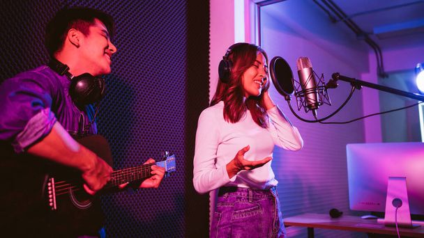 Pretty Asian female singer recording songs by using a studio microphone and pop shield on mic with male playing guitar in blue and red light. Performance and show in the music business. Duet session. - Фото, зображення