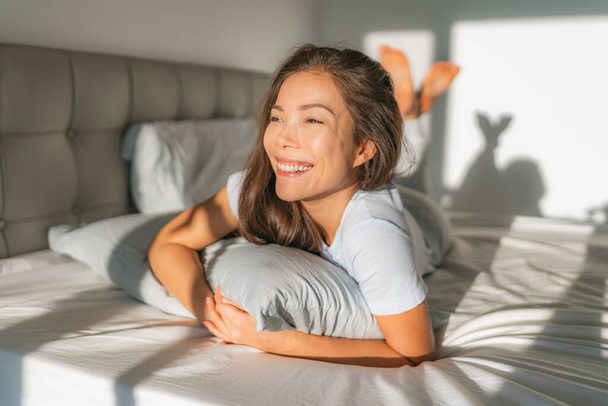 Bed woman waking up early morning happy enjoying sun on comfortable mattress and pillow. Asian girl relaxing in bedroom smiling. Natural beauty after healthy good night sleep sleeping in on weekend - Zdjęcie, obraz