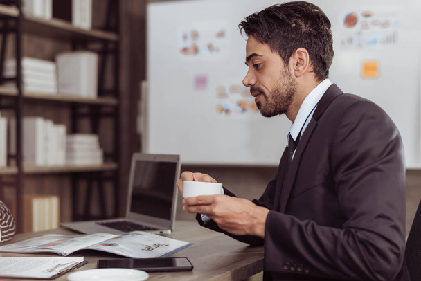 Young smart businessman drinking coffee in working room. Handsome arabic businessman sitting and holding cup of coffee. Business and working concept. - 写真・画像
