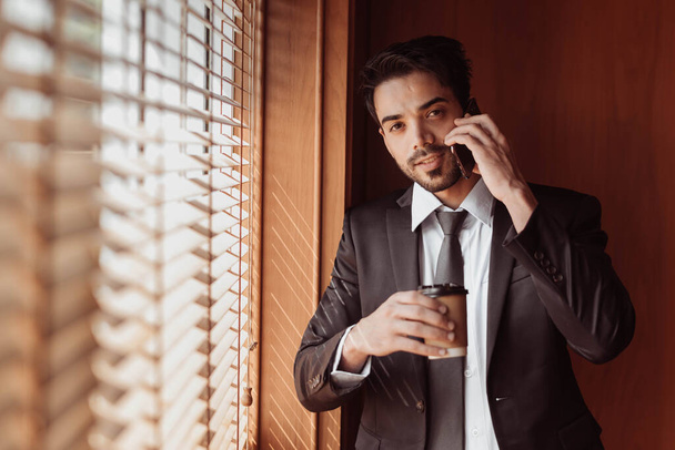 Young businessman using smartphone and holding cup of coffee in working room. Handsome arabic businessman in black suit talking on smartphone. - Photo, Image