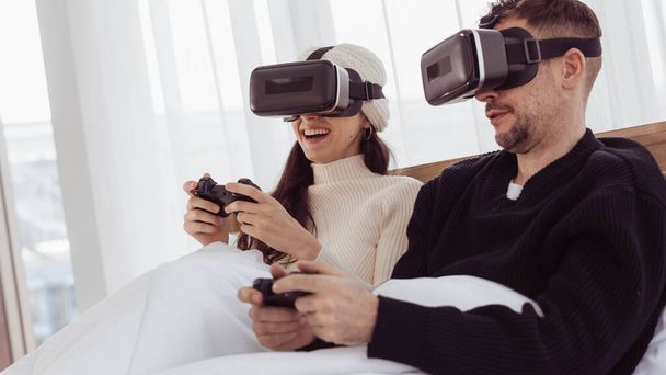 Любящая пара в очках играет в видеоигру с джойстиками на кровати. Счастливые муж и жена носят очки виртуальной реальности и вместе играют в видеоигры дома. - Фото, изображение