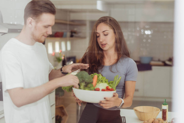 Šťastný pár rád vaří spolu v kuchyni. Mladý milující muž a žena stojící a držící vegatable v bílé kuchyni pokoj. - Fotografie, Obrázek