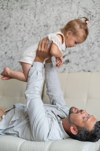 A Caucasian man lies on the couch and holds a little daughter above him. Caring loving dad raised the child up. Happy fatherhood. - Foto, afbeelding