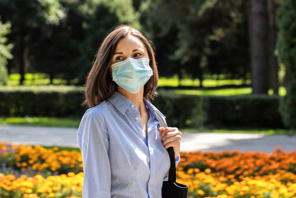 Beautiful young woman walking in a summer park wearing a protective medical mask - Фото, изображение