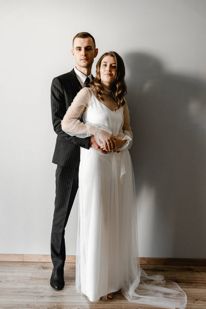 a wedding couple stands near a plain wall. beautiful wedding clothes. bride holding a bouquet of flowers. young family. happy people. joyful moment. - Valokuva, kuva