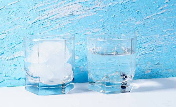 Дві склянки, одна склянка повна льоду, інша половина з водою на синьому фоні. Концепція танення льоду
. - Фото, зображення