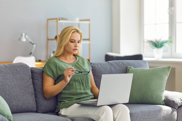 Focused freelancer woman working on laptop sitting on sofa in living room of her home. - Фото, зображення
