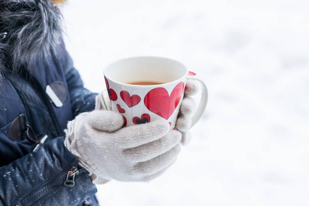 Женщина пьет горячий напиток. Чашка чая с красными сердцами. Руки женщины в белых вязаных варежках, керамическая кружка с кофе или чаем на фоне белого снега. Пикник на открытом воздухе на день святого Валентина. - Фото, изображение