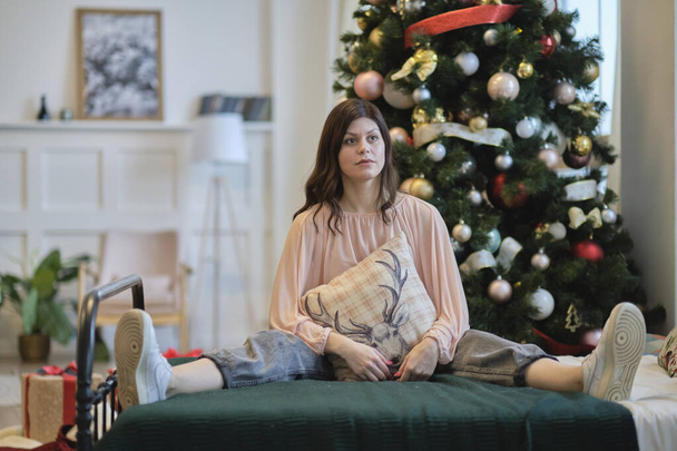 fiatal nő ül az ágyon háttal egy karácsonyfa. az élet újrakezdésének koncepciója az ünnepségek után. - Fotó, kép
