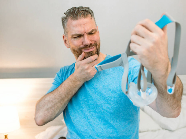 Unhappy shocked man with chronic breathing issues surprised by using  CPAP machine sitting on the bed in bedroom. Healthcare, CPAP, Obstructive sleep apnea therapy, snoring concept - Photo, Image