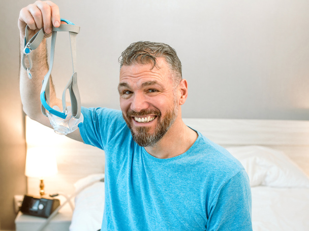 Mature happy man with chronic breathing issues considers using sitting on the bed in bedroom with CPAP machine. Healthcare, CPAP, snoring concept - Photo, Image