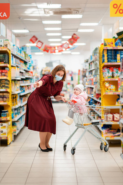 New normal. A young mother with a medical mask on her face shows something with her hand to her baby sitting in a grocery cart. The concept of shopping during the coronavirus pandemic. - Photo, Image