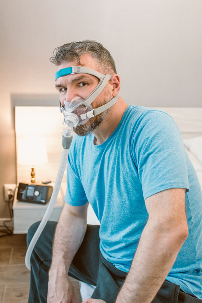 Dojrzały szczęśliwy człowiek z chronicznymi problemami z oddychaniem przy użyciu CPAP maszyna siedzi na łóżku w sypialni. Opieka zdrowotna, CPAP, koncepcja chrapania - Zdjęcie, obraz