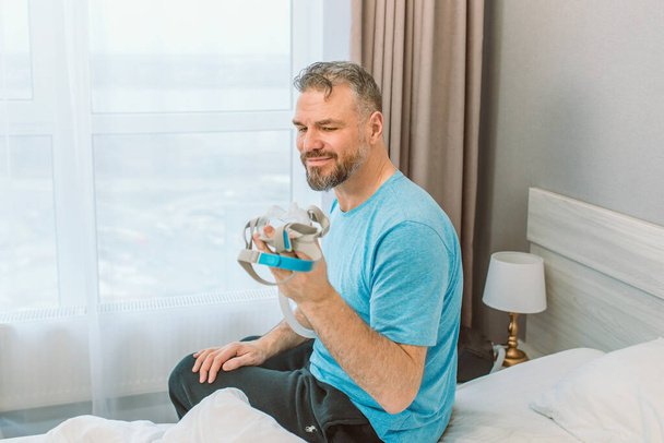 Uomo maturo felice con problemi respiratori cronici considera sedersi sul letto in camera da letto con macchina CPAP. Sanità, CPAP, russare concetto - Foto, immagini