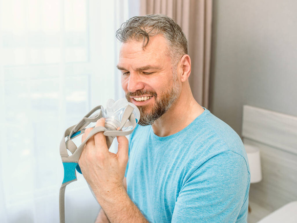 Homme heureux mature avec des problèmes respiratoires chroniques envisage d'utiliser la machine CPAP assis sur le lit dans la chambre. Soins de santé, CPAP, concept de ronflement - Photo, image