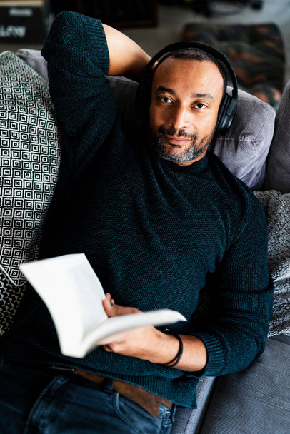Jóképű férfi otthon pihen a kanapén egy könyvvel és fejhallgatóval. - Fotó, kép