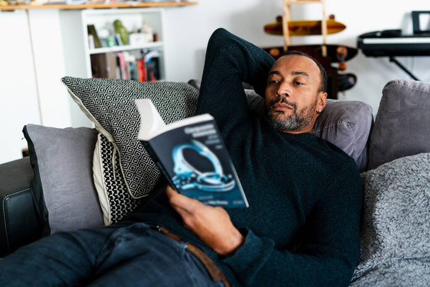 Smíšené rasy muž ležící na pohovce doma a čtení knihy - Fotografie, Obrázek