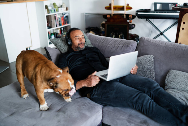 40 years old mixed-race man relaxing on the sofa and watching a movie next to his dog - Photo, Image