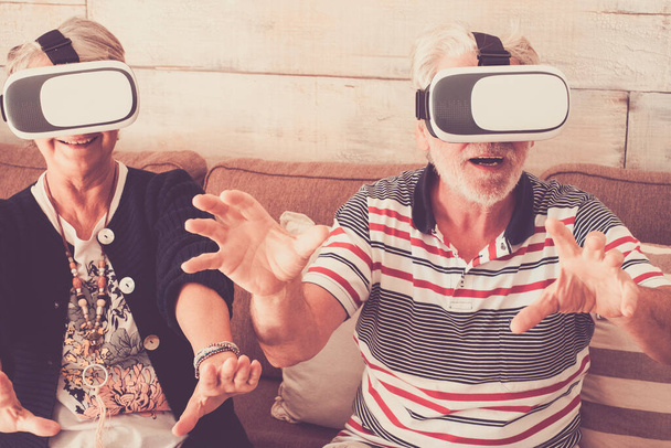 Νεανικό concept με το ηλικιωμένο ζευγάρι να απολαμβάνει goggled ακουστικά μαζί σε online μοντέρνα παιχνίδια και εμπειρία εικονικής πραγματικότητας  - Φωτογραφία, εικόνα