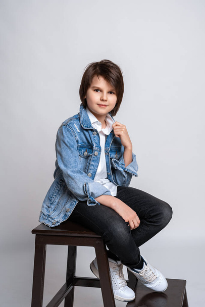 Handsome fashion little boy 11-13 years old sits on a chair in studio. Dressed in a white shirt, black jeans, jean jacket and white sneakers. Model Test. Fashion and people concept - 写真・画像