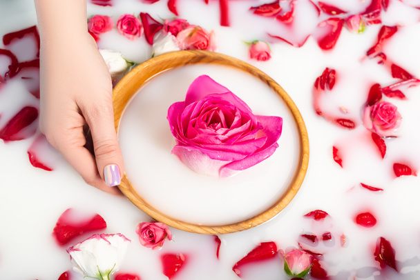 cropped view of woman holding wooden bowl with pink peony near roses in milky water  - Foto, afbeelding