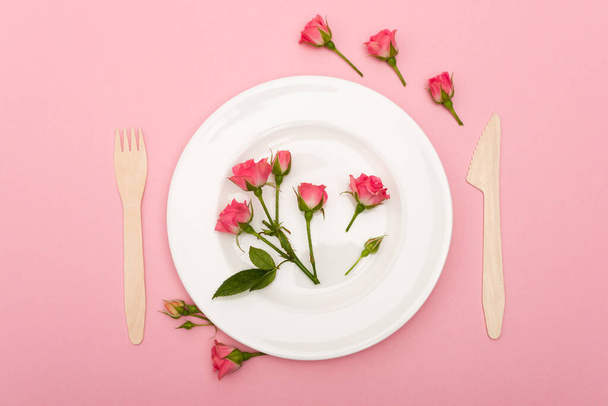 vista superior de cubiertos de madera desechables cerca de plato blanco con flores aisladas en rosa  - Foto, Imagen
