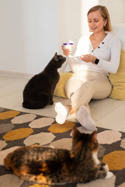 Mulher alimenta um gato com iogurte de uma colher, sentado no chão. Vida em casa com seus animais de estimação - Foto, Imagem