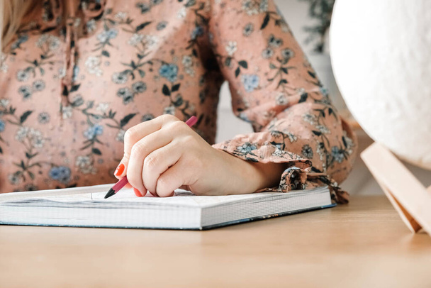 Vrouw handen met potlood, ze is aan het schrijven in haar notitieboekje zittend aan de tafel met boekenplank achtergrond. Kopieer, lege ruimte voor tekst. - Foto, afbeelding