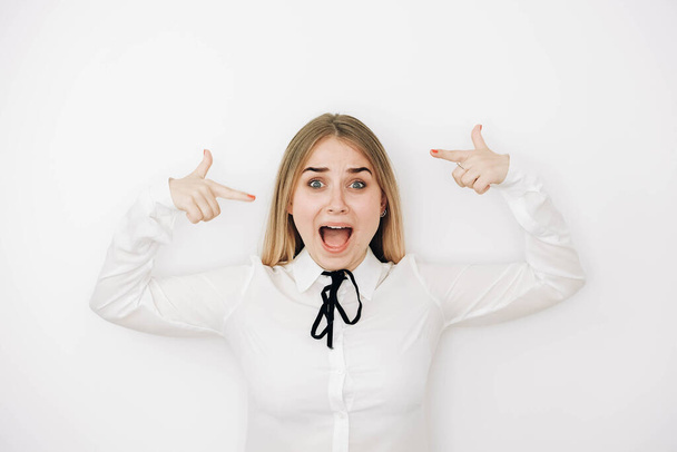Beautiful blonde woman wearing stylish white shirt over white background amazed and surprised looking and pointing with fingers and raised arms.Copy, empty space for text. - Photo, Image