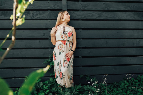 Beautiful blonde woman dressed in flower dress in a green leaves trees on a wooden wall background. Copy, empty space for text. - 写真・画像