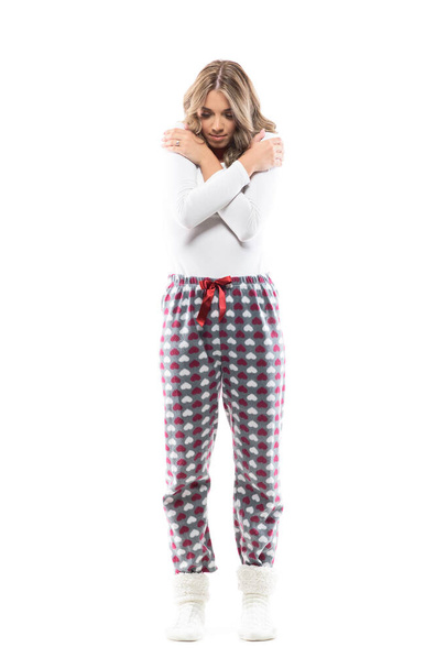 Cozy young beautiful woman in pajama and winter socks embracing herself looking down feeling cold. Full length portrait on white background. - Valokuva, kuva