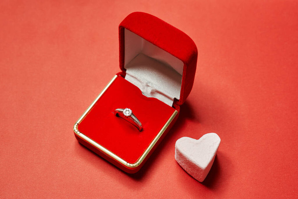на червоному тлі червона коробка з обручкою і зефірним серцем. пропозиція одружитися в день валентина
                            - Фото, зображення