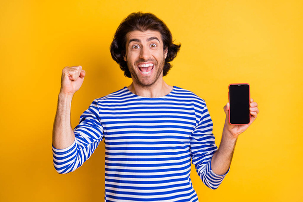 Photo of crazy ecstatic guy hold smartphone raise fist wear nautical vest isolated over shine yellow color background - Foto, imagen