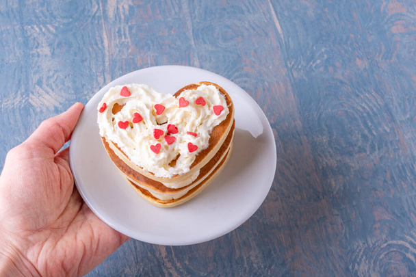 Female hand holding a white plate with a stack of homemade heart shaped pancakes decorated with white cream with red hearts on a blue wooden background - Fotografie, Obrázek