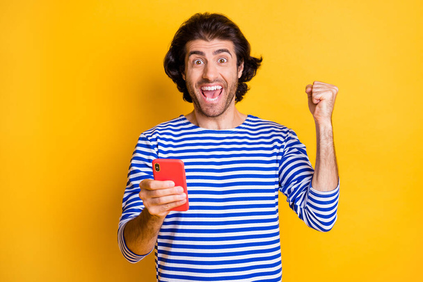 Photo of ecstatic man hold smartphone raise fists scream wear blue white shirt isolated on shine yellow color background - Photo, image