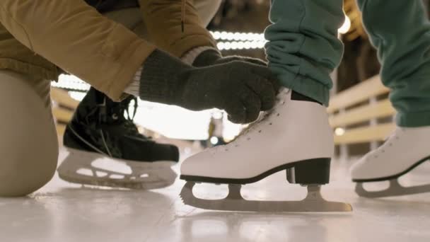 Низкочастотный крупный план неузнаваемых мужских рук в зимних перчатках, завязывающих шнурки на белых фигурных скитальцах на открытом катке вечером - Кадры, видео