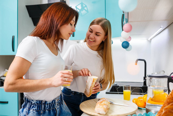 dwie piękne młode kobiety przyjaciele stoją przy stole w kuchni w domu i jeść chleb i pić sok pomarańczowy - Zdjęcie, obraz