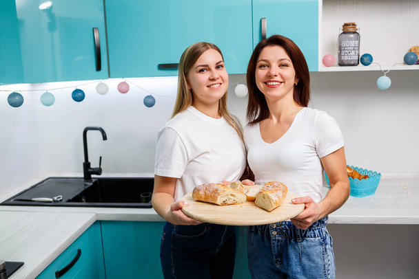 two beautiful young women friends stand at the kitchen table at home and eat bread and drink orange juice - Fotoğraf, Görsel