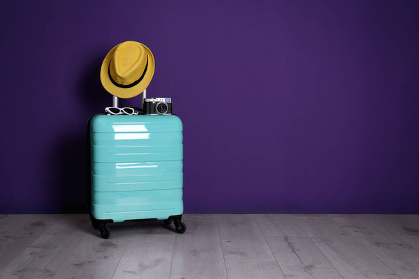 Travel suitcase with hat, camera and sunglasses on wooden floor near purple wall, space for text. Summer vacation - Foto, Bild