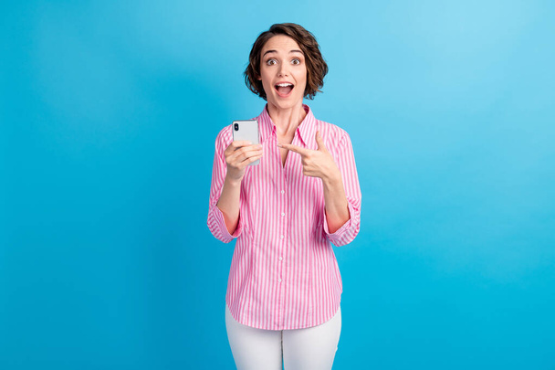 Фото шокированной удивленной девушки указывать пальцем смартфон носить розовый белый брюки изолированы на синем фоне цвета - Фото, изображение